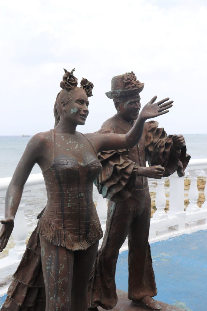 A year-round bronze sculpture of a king and queen of an early Carnaval on Cozumel. The woman is wearing a fancy long gown and has her hair in an up-do. The man has an embellished hat and his shirt has wide full ruffled sleeves. A white railing along the malecon is seen in the background and then the ocean beyond. The sculpture appears weathered with age.