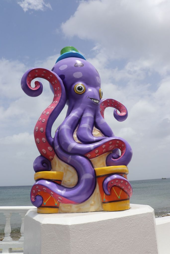 A whimsical cartoon octopus sculpture is resting on the white cement seawall. This decoration for Carnaval season in Cozumel Mexico is purple and orange with a gold base. The ocean and cloudy sky are visible in the background.