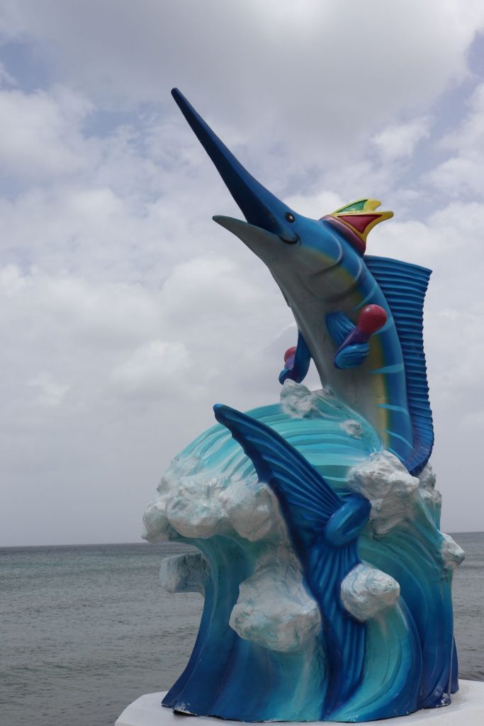 A swordfish sculpture that is part of the Cozumel Mexico Carnaval season along the malecon. The ocean is visible behind. The decoration is whimsical with a crown on the swordfish's head and purple maracas held in his fins. He is posed on a pedestal that looks like ocean waves with light blue, medium blue and white foam.