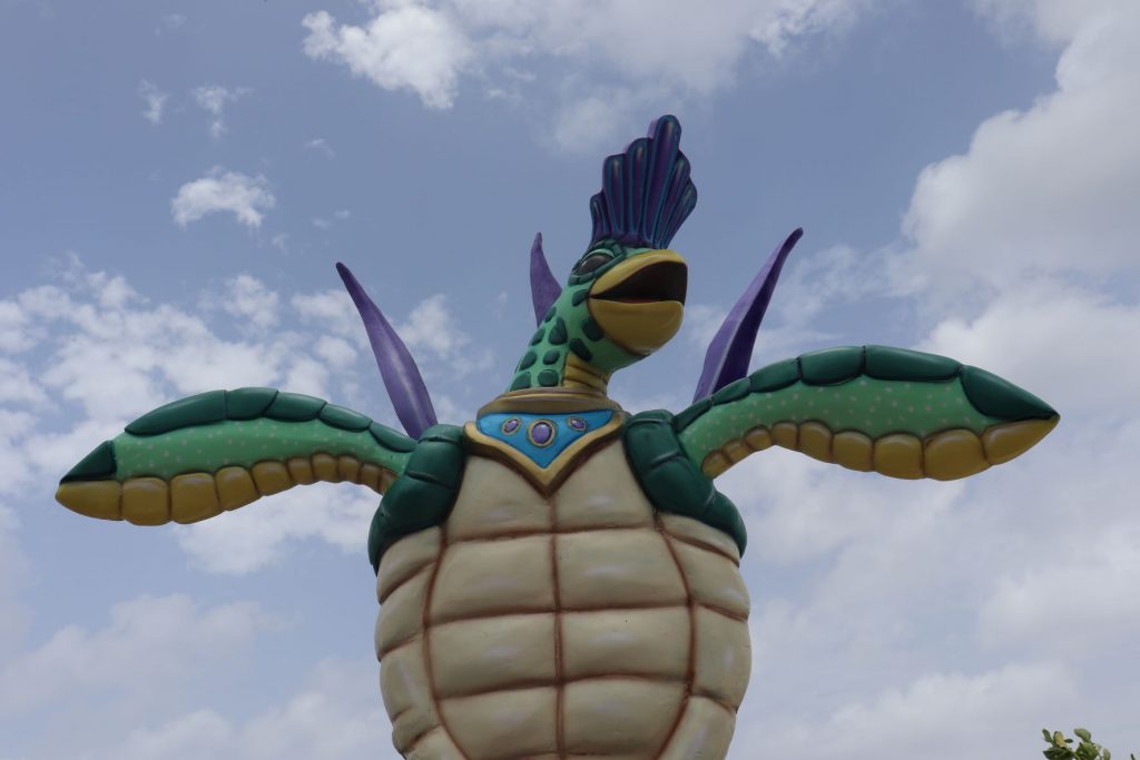 A brightly colored whimsical sea turtle sculpture is part of the Cozumel Mexico decoration for Carnaval season. The turtle is mostly blue and green with a purple festival headpiece and tail feathers, as if in costume. There is a cloudy sky above.
