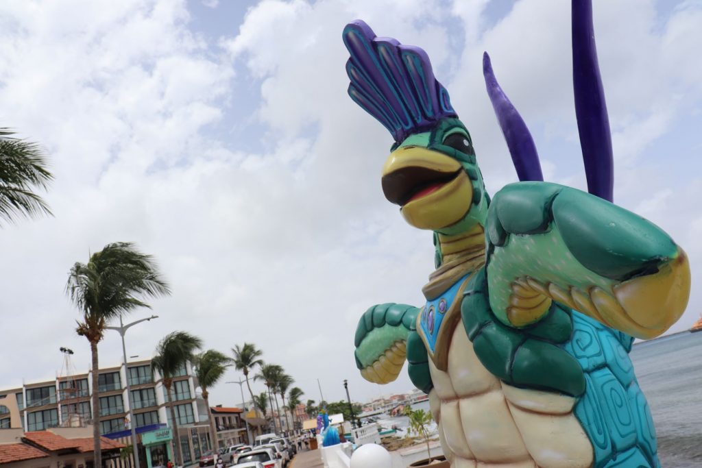 A brightly colored whimsical sea turtle sculpture is part of the Cozumel Mexico decoration for Carnaval season. The turtle is mostly blue and green with a purple festival headpiece and tail feathers, as if in costume. There is a cloudy sky above and a view of the main street with small hotels, shops and parked cars. Tall palm trees line the street median strip. You can see the ocean to the far right.