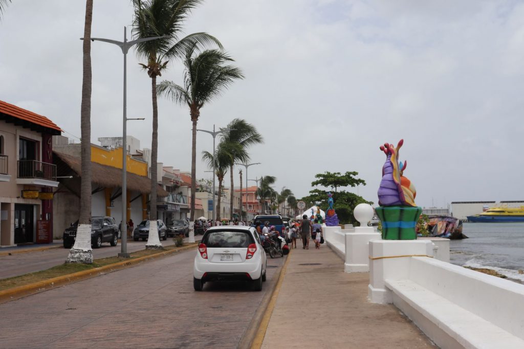 A view of the main street along the ocean in Cozumel Mexico at Carnaval season. There are shops and businesses along the left side,, with a tall palm lined median and the malecon with a white seawalll along the right. There are bright whimsical sea creature sculptures on white cement pedestals as part of the sea wall. The closest one is a purple, gold and orange sea snail. There is a ferry waiting at the ferry terminal in the distance.