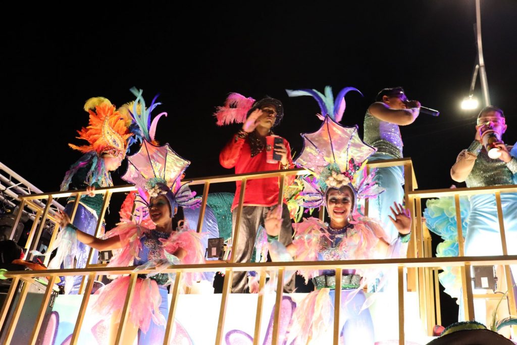 Two young women wearing costumes of white dresses heavily embellished with fluffy white feathers and tall feathered headpieces wave to spectators as they ride on a parade float for Cozumel Carnaval.