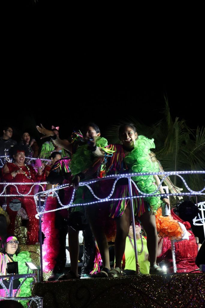 With dark black sky as background, three young women with large lime green ruffled boas ride on a Cozumel Carnaval parade float.