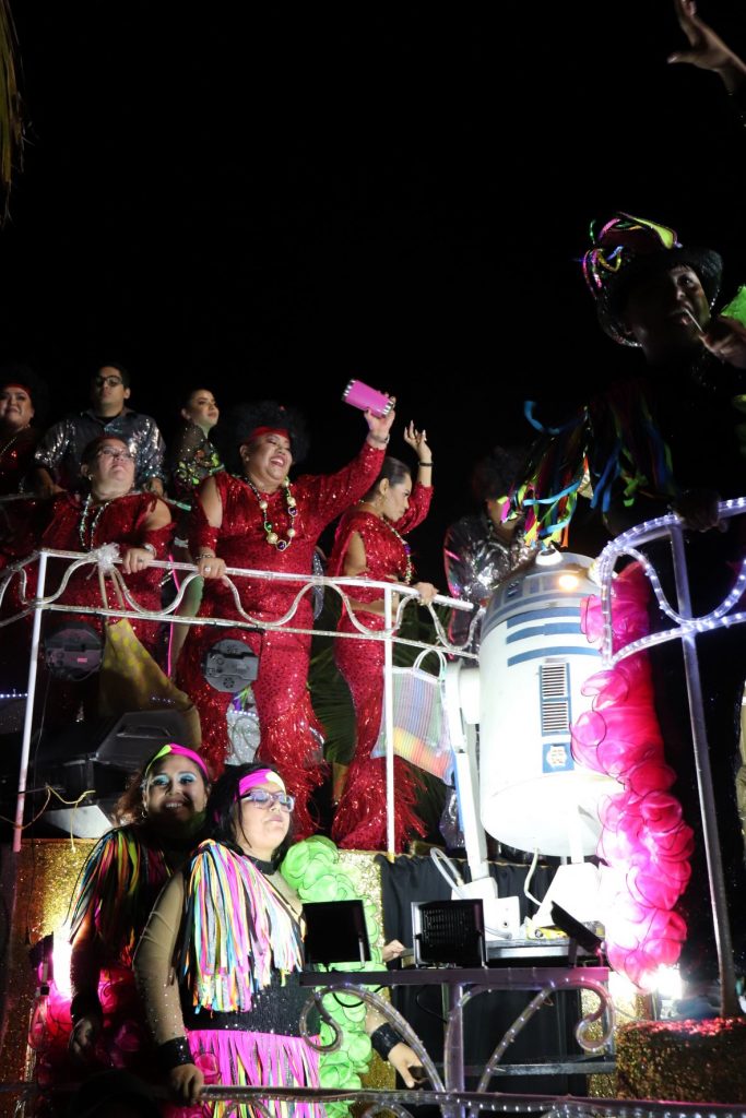 A parade float that features a replica of R2D2 robot character from Star Wars. There are entertainers riding on the float and smiling as they toss beads and cups into the crowds. Three womennare in long red gowns