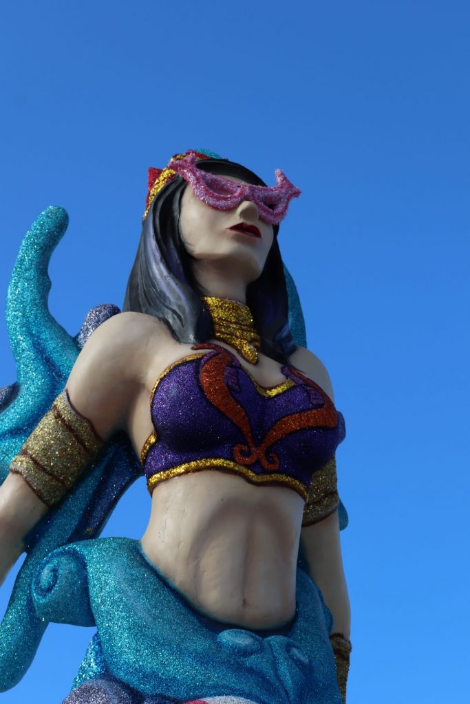 Mermaid decoration along the malecon in Cozumel, Mexico for Carnaval. This larger than life-size sculpture is whimsical with gold glittery arm bands and purple glitter bustier, caucasian skintone, dark long hair and fuschia glitter sunglasses and aqua backpiece. The lower portion is dark and medium blues. A clear blue sky is seen behind the mermaid.