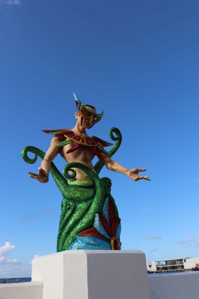 Mermaid decoration along the malecon in Cozumel, Mexico for Carnaval. This larger than life-size sculpture is whimsical with red glittery arm bands and bustier, caucasian skintone, dark long hair and fuschia glitter crown and backpiece. The lower portion is dark and medium blues with a gold and red glitter star in the center. A clear blue sky is seen behind the subject. The mermaid has her hands outstretched in a welcoming gesture.