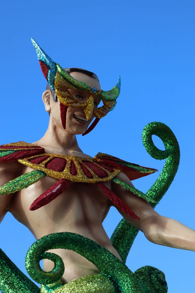 Merman decoration along the malecon in Cozumel, Mexico for Carnaval. This larger than life-size sculpture is whimsical with red and gold glittery yoke, green glittery tendrils and a carnaval style mask with aqua, purple, gold and green glitter. A clear blue sky is seen behind the subject.