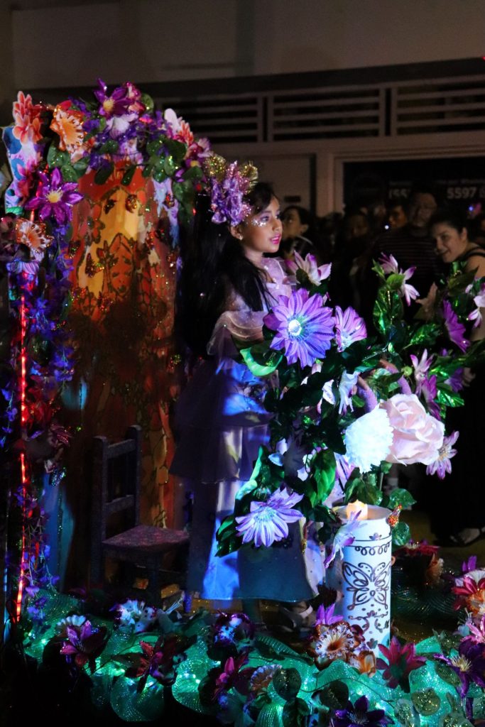 A young woman smiling as she rides on a flower filled parade float as part of Cozumel Carnaval. She has long dark hair, and the flowers obstruct the view of her costume.