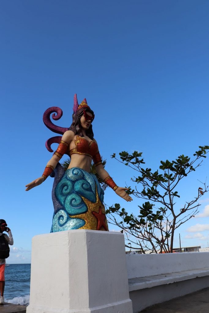 Mermaid decoration along the malecon in Cozumel, Mexico for Carnaval. This larger than life-size sculpture is whimsical with red glittery arm bands and bustier, caucasian skintone, dark long hair and fuschia glitter crown and backpiece. The lower portion is dark and medium blues with a gold and red glitter star in the center. A clear blue sky is seen behind the subject. The mermaid has her hands outstretched in a welcoming gesture.