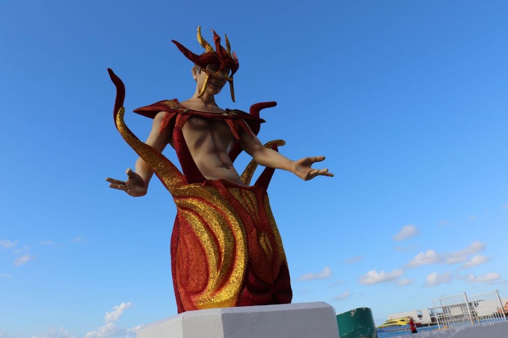 The full length of a merman decorative sculpture for Carnaval is seen mounted on a pedestal base that is part of the wall along the malecon in Cozumel Mexico. His costume reveals his bare muscular and trim torso with red and gold glitter tendrilled base, yoke and mask. His arms are outstretched in a welcoming gesture. There is a ferry at the dock in the distance` and clear blue skies.
