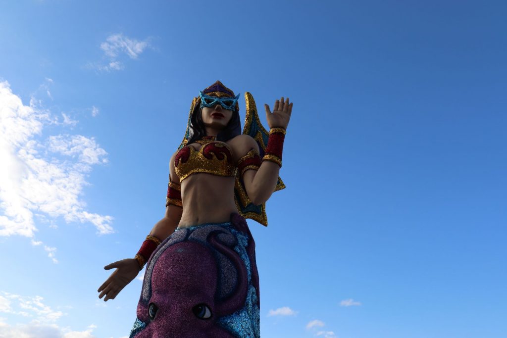 Mermaid decoration along the malecon in Cozumel, Mexico for Carnaval. This larger than life-size sculpture is whimsical with fuschia glittery arm bands and bustier, caucasian skintone, dark long hair and fuschia with gold glitter crown and backpiece. The lower portion is dark and medium blues with a purple octopus in the center. A clear blue sky is seen behind the subject. The mermaid has her hands outstretched in a welcoming gesture. She is wearing aqua glitter sunglasses.