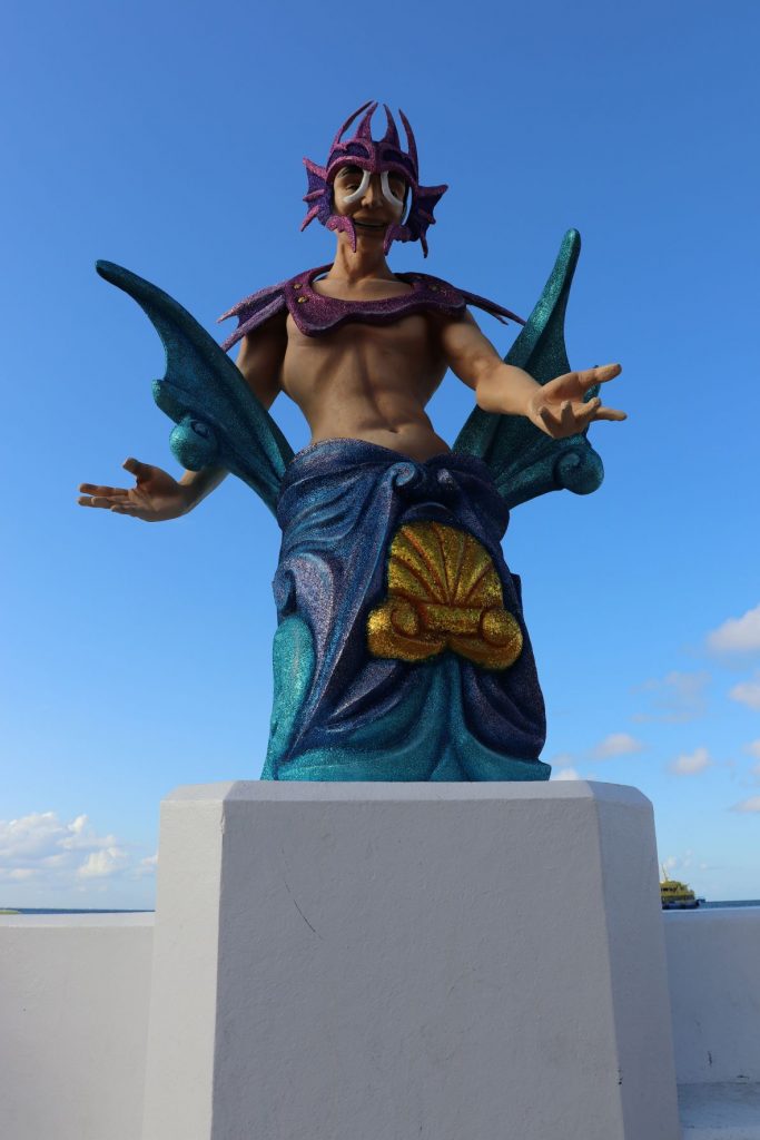 A full length view of a merman sculpture decoration that is mounted on a pedestral that is part of the wall along the malecon in Cozumel Mexico. The wall and pedestal are white cement. The lower portion of the merman is aqua, blue and purple and evokes images of waves with a gold seashell motif in the center. The merman's torso is trim and muscular with caucasian skintone. His arms are outstretched in a showmanship manner. He has a fuschia and purple glitter yoke and headpiece. The background is a clear blue sky.