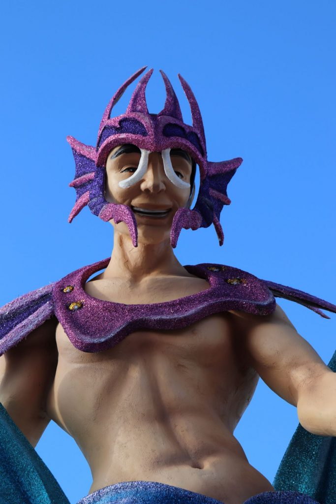 Merman decoration along the malecon in Cozumel, Mexico for Carnaval. This larger than life-size sculpture is whimsical with a fuschia and purple glitter headpiece and yoke,, over a sculpted muscular yet trim,, cuacasian torso. The sky behind the merman is crystal clear.