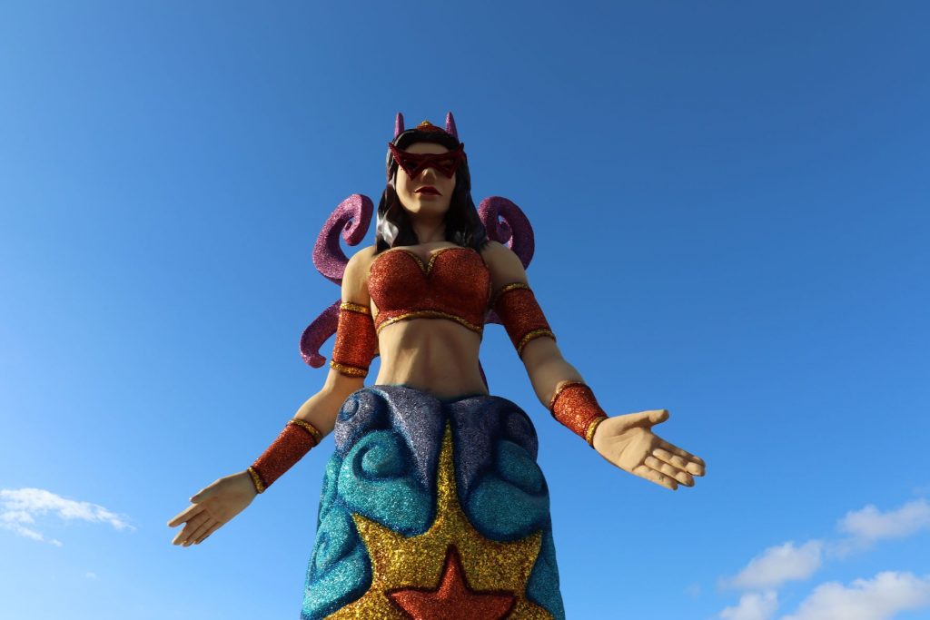 Mermaid decoration along the malecon in Cozumel, Mexico for Carnaval. This larger than life-size sculpture is whimsical with red glittery arm bands and bustier, caucasian skintone, dark long hair and fuschia glitter crown and backpiece. The lower portion is dark and medium blues with a gold and red glitter star in the center. A clear blue sky is seen behind the subject. The mermaid has her hands outstretched in a welcoming gesture.