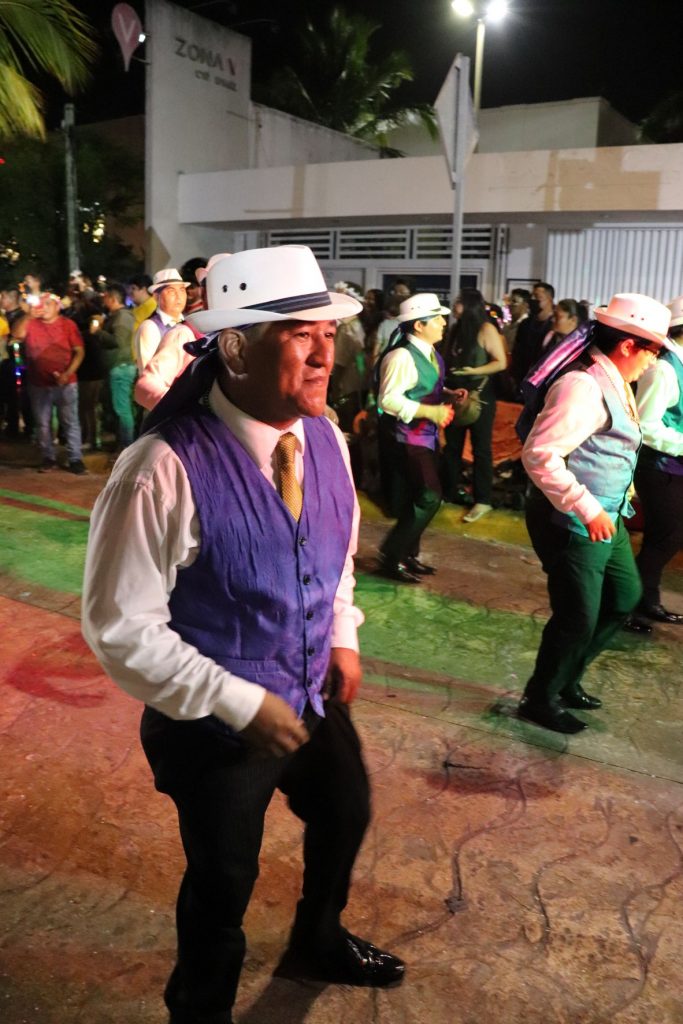 A middle-aged latino man performing as part of a large latino dance troupe. They are wearing dark slacks, long sleeve white shirts, gold ties, purple vests and white fedoras. These older men dance the entire length of the Cozumel Carnaval parade each night. This photo has 4 other members in the background.