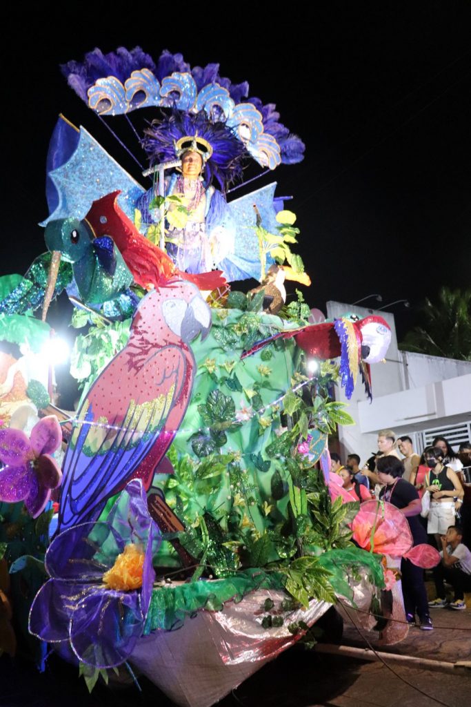 A Cozumel Carnaval parade float decorated with lush green foliage and a 5 foot tall red parrot. An entertainer rides on the very top with a large purple fan designed backpiece and a purple feathered headpiece.