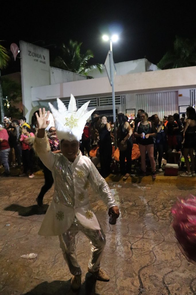 Male entertainer walking and waving in one of Cozumel Carnaval parades. He is dressed in all white with metallic gold trim on his tunic. He has a large white and gold headpiece shaped like a 18" tall crown.