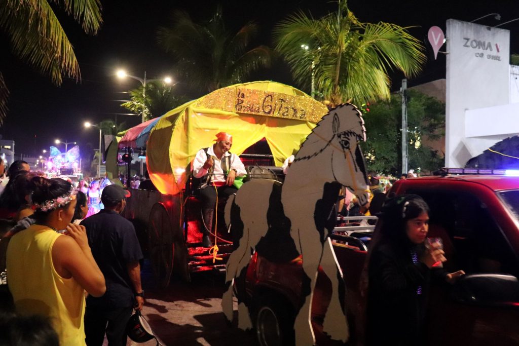 A bright yellow gypsy wagon with a costumed character driver being pulled by a red pickup truck in one of the Cozumel Carnaval parades. The pickup truck had life size black and white painted horses attached to the sides,, so it appears like the horses are pulling the gypsy wagon.