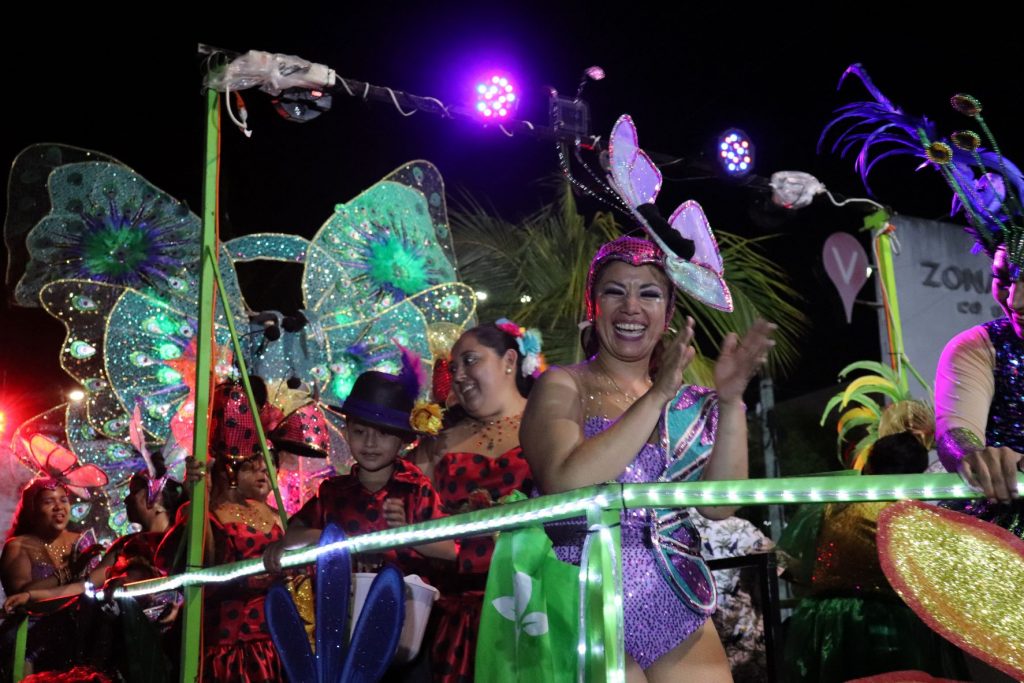 A Butterfly themed parade float as part of Cozumel Carnaval. Several women are riding the float in ladybug themed costumes that are red and black. The woman in the front is clapping and smiling at spectators. She is wearing a purple butterfly themed costume.