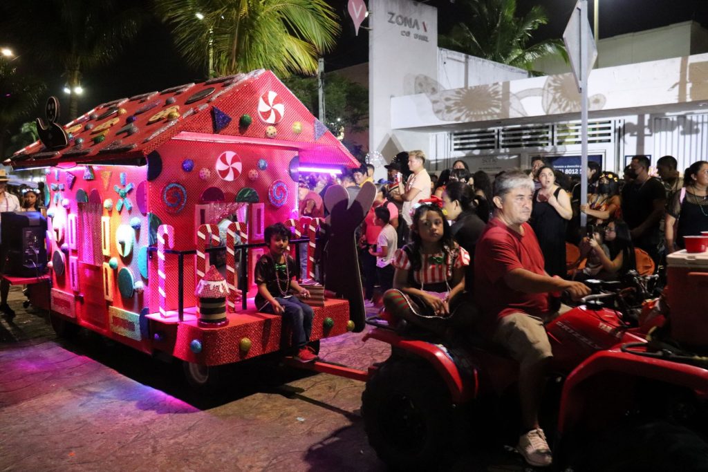 Two men riding on a red ATV pulling a Christmas candy house as a float behind them, during a Cozumel Carnaval parade.