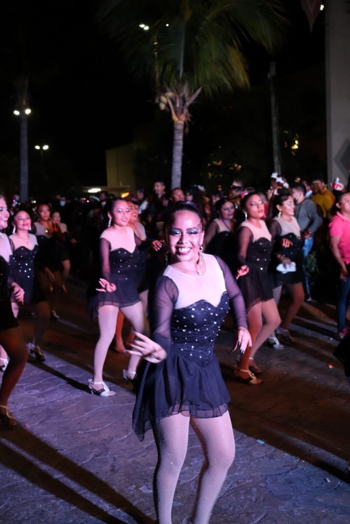 Closeup of a young latino woman who is part of a dance troupe dancing and walking in one of the Cozumel Carnaval parades. She is wearing a short black sparkly costume dress with sheer black lomg sleeves and a large white yoke. Other members in matching outfits follow behind her. You can see spectators in the darkness along the street.