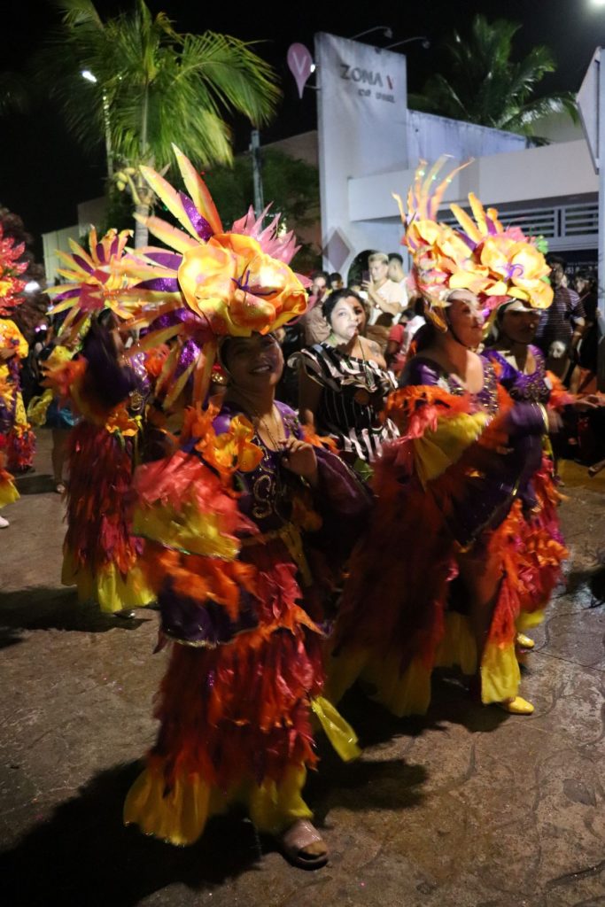 Bright orange, yellow and purple feathered costumes on two entertainers walking in the Cozumel Carnaval parade. Others follow behind and there are spectators lining the street.