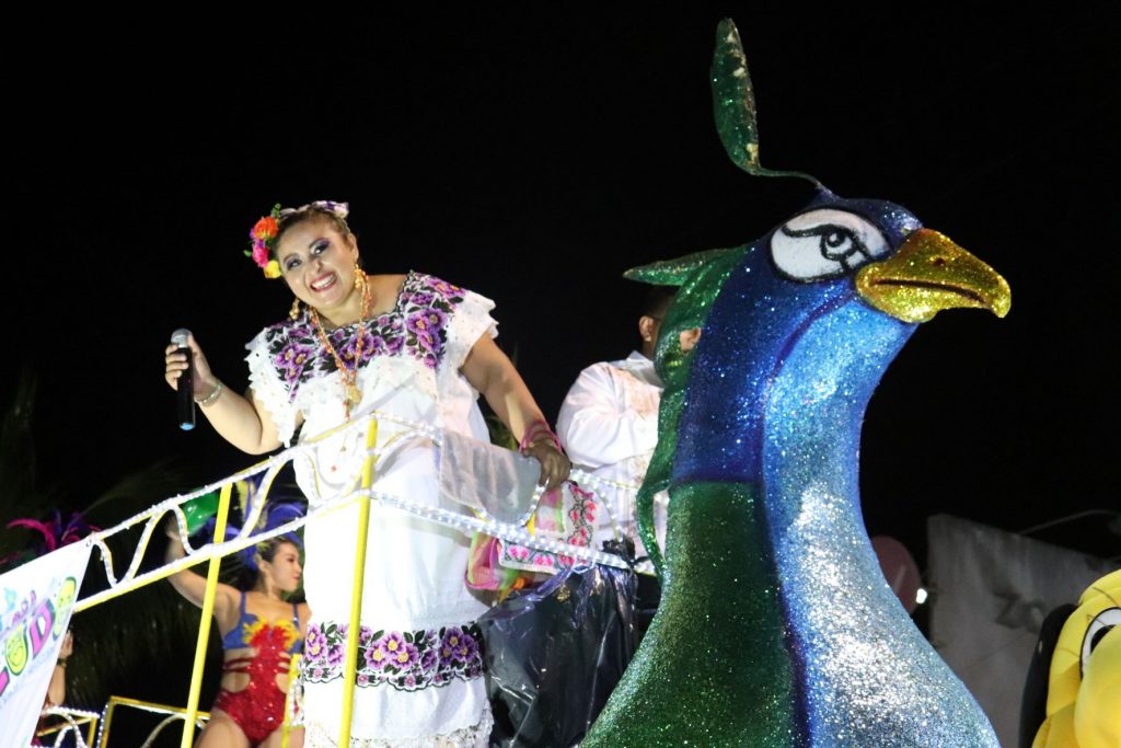 A Mexican woman holding a microphone and wearing a traditional embroidered dress is riding on a parade float and leans over smiling into the camera. This parade float features a large purple and green bird. There are other costumed performers in the background. The background is night darkness as this Cozumel Carnaval parade is held.