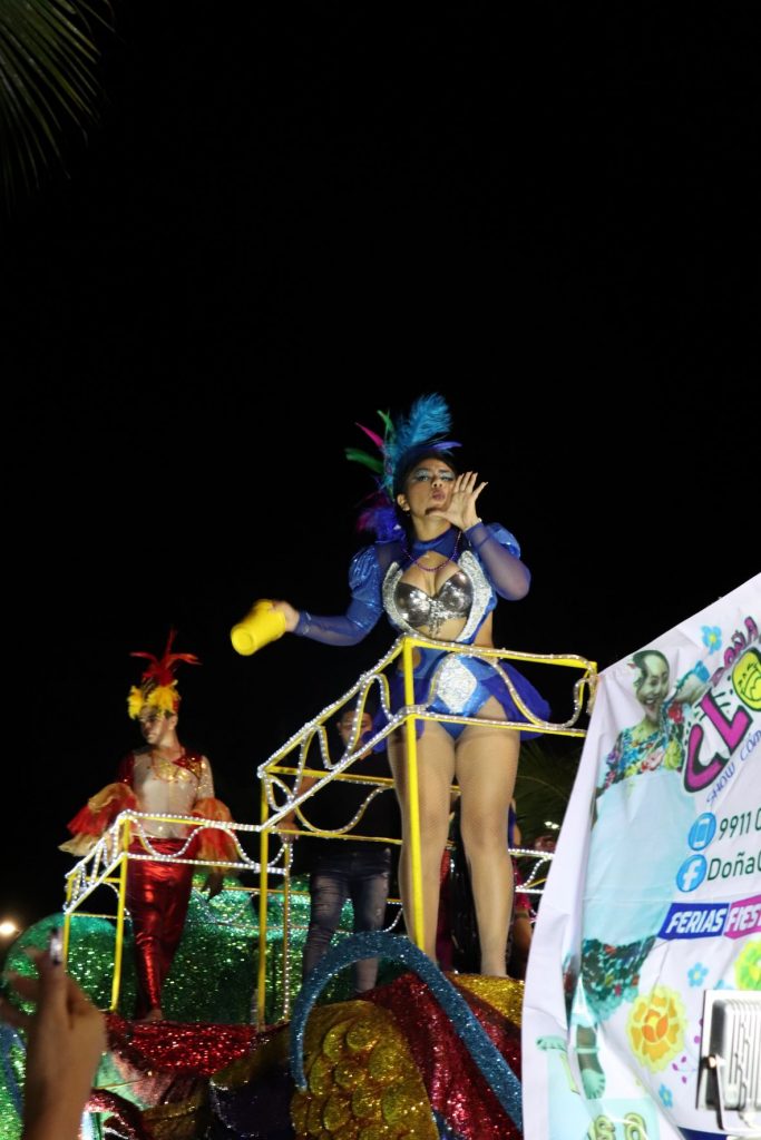 A parade float from Cozumel Carnaval with a young woman wearing a purple and silver costume. She has her hand positioned by her mouth to shout loudly and in the other hand she has a yellow cup to toss to the crowd. Behind her is a young man wearing a bright red and silver outfit while riding on another area of the float.