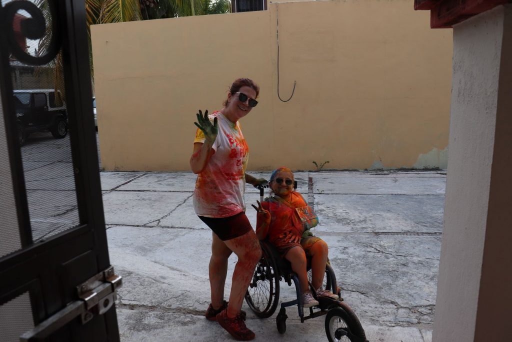 Anna and her mom smile and raise their hands to wave "hi" at the camera. They are both covered head to toe with dusty paint. Anna's wheelchair and FreeWheel is equally messy.