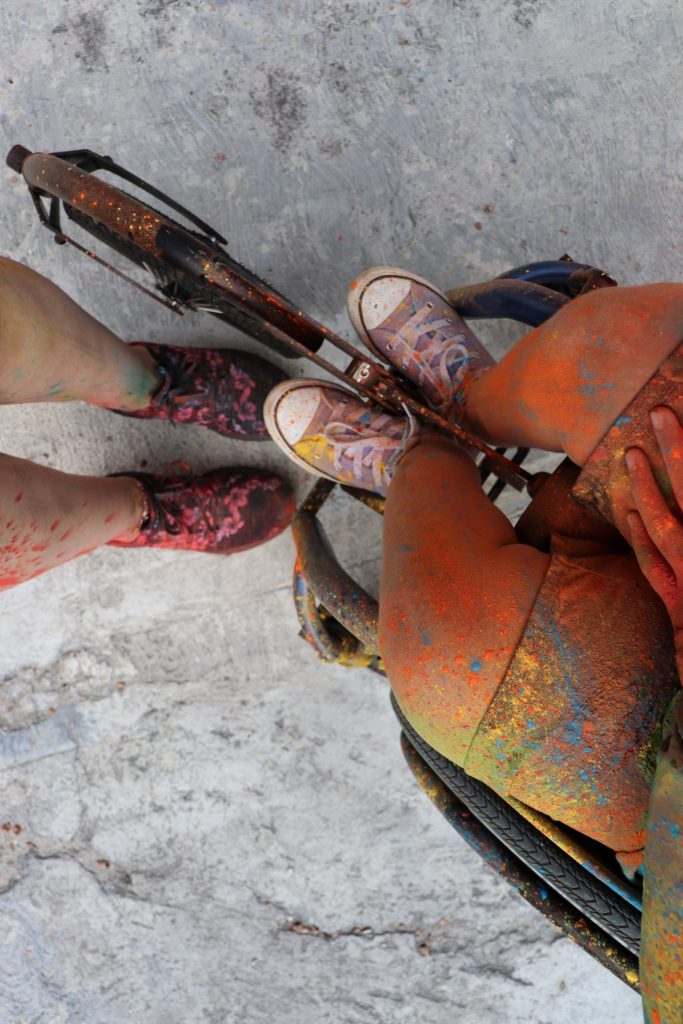 A photo looking down at two pairs of colorful shoes is seen. They are covered with paint of all colors. Anna, a teenage girl with a wheelchair is wearing purple shoes. Her blue wheelchair has the paint everywhere on it.