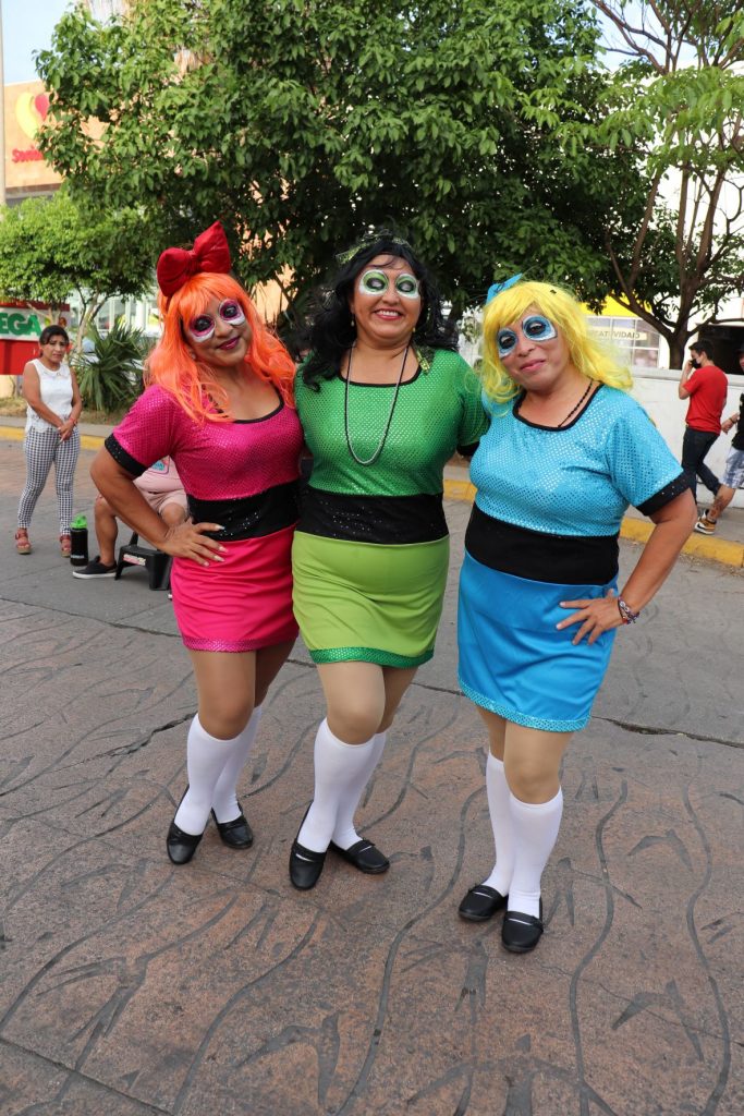 Three women dressed in similar dresses and matching white knee high socks with black shoes. They are wearing bright long hair wigs and exaggerated eye makeup. One dress is hot pink, another is lime green and the final one is light blue. They pose arm in arm on the street before one of the Cozumel Carnaval parades begin.