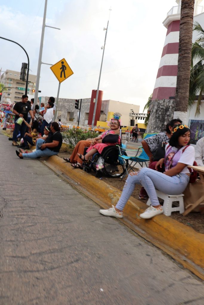Spectators gathered along the Cozumel Carnaval parade route before the event begins. A teen girl, Anna, sits up on the sidewalk in her wheelchair for an accessible view of the parade.