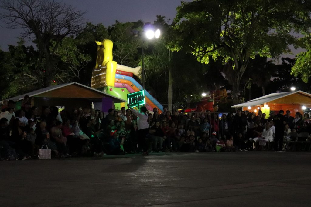 A seated and standing room crowd lines the stage area waiting for Carnaval dance competitions at Parque Quintana Roo in Cozumel Mexico. You can see children's amusement rides and vendor huts in the background.