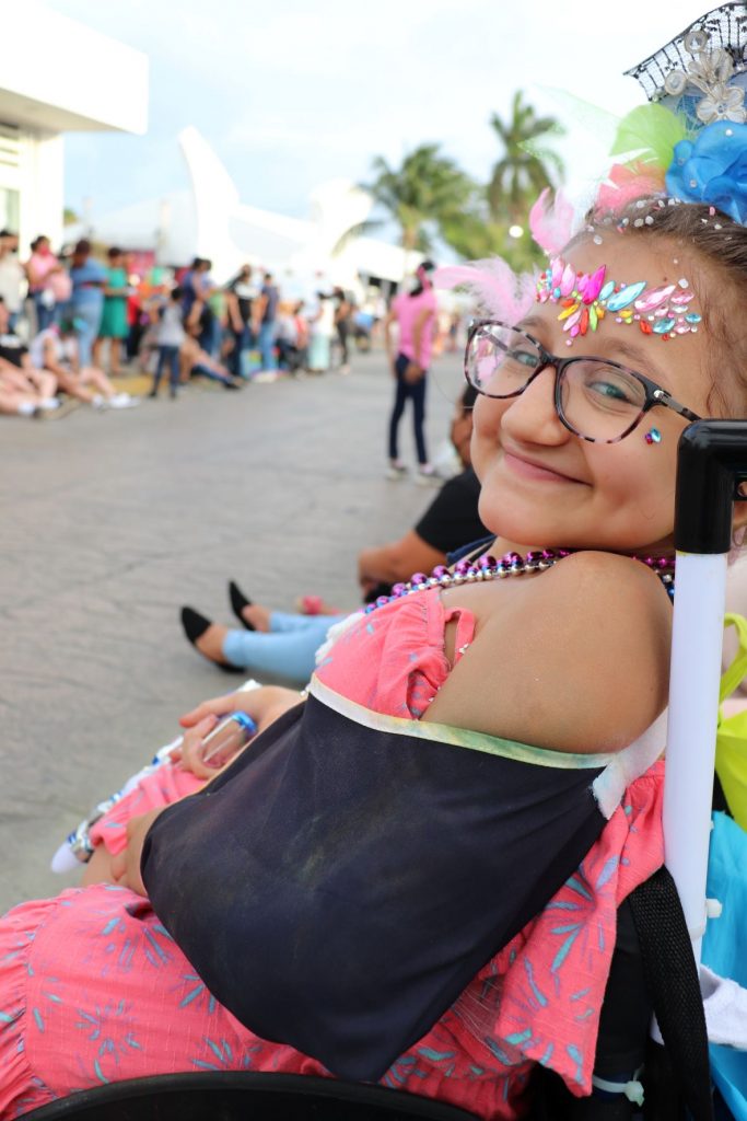 A teen girl, Anna, wearing a pink dress with gems on her forehead sits in a wheelchair about to enjoy the parades of Cozumel's carnaval celebration showing the accessibility. She has an unobstructed view.