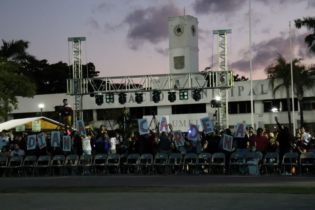 Seating for dance competitions in front of the municipal palace of Cozumel Mexico. There is scaffolding rigged with lighting and a queen candidate's fans have assembled with large placards spelling out her name to encourage votes for this queen candidate. The chairs are standard folding chairs in a row and the building is white, against a dusky cloudy sky.