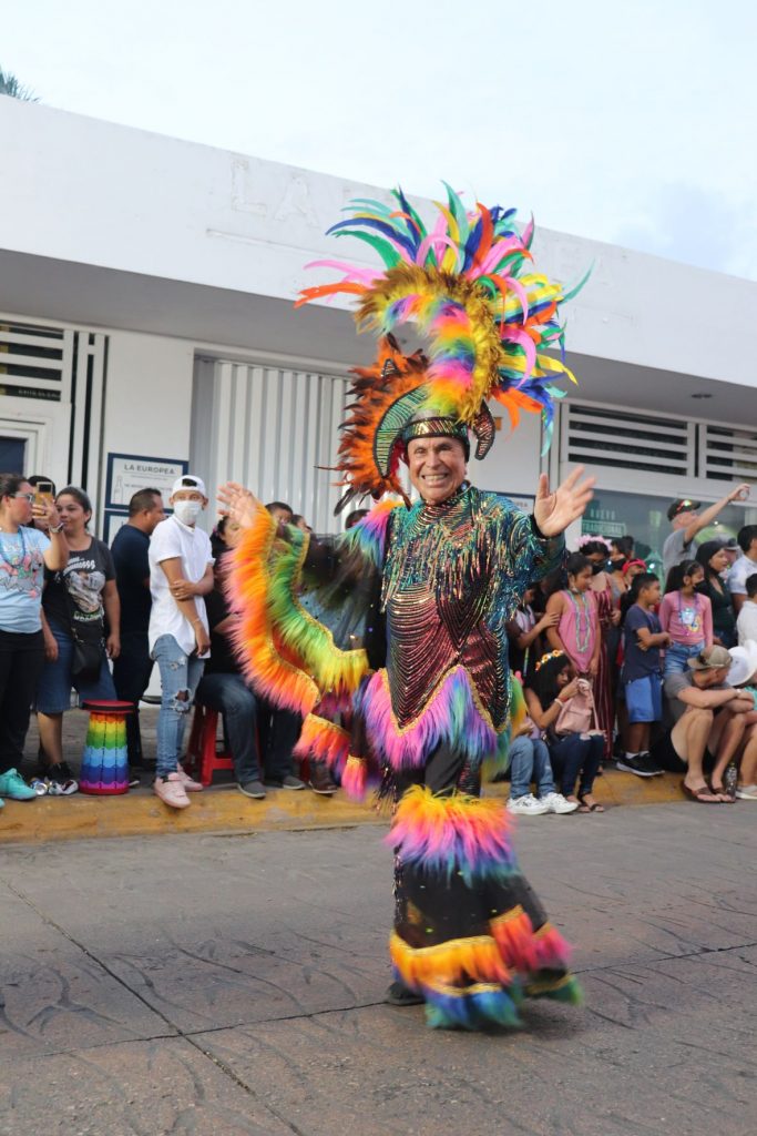 Mr. Carnaval, also known as Mr. Spectacular Fernando Ferraez serves as the grand marshal and emcee for many carnival events. He is known for outlandish outfits. This is him walking in one of the Cozumel Carnaval parades wearing a flamboyant black outfit and waving to the crowd as he passes. He has a very tall headpiece with assorted bright feathers and his outfit has many layers of fringe and feathers that are yellow, orange, pink, blue and green.