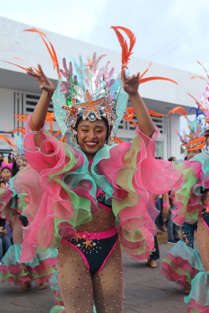 This young woman is part of a dance troop performing in one of the Cozumel Carnaval parades. She is wearing a black and pink swimwear styled bikini with sheer stockings that have gold sparkle gems attached. Her arms are both raised in the air and are covered with large multi color ruffles. She is smiling and has a headpiece with an ocean theme. There are stylized coral and starfish along with tall spikey orange feathers. It is still daylight and a white building is visible in the background.