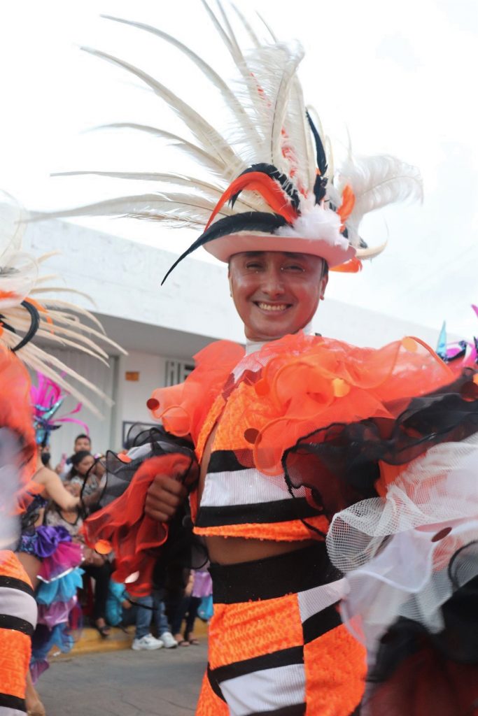 A closeup photo of a young latino man wearing a bright orange and white costume with very full ruffled sleeves. He has a white fedora with tall spikey white feathers and a few decoartive orange and black smaller feathers in the band. He is marching in one of the Cozumel Carnaval parades, with spectators lining the street.