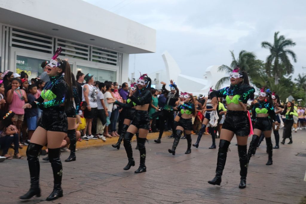 A dance team performing and marching in one of the Cozumel Carnaval parades. The young women are wearling silver upper face masks with pink feathers above the masks. They have hot lime green bra tops, black short shorts and black over the knee boots. It is daylight and there are spectators and a white building in the background.