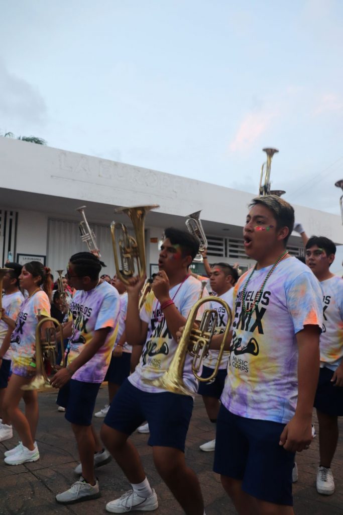 Two rows of the marching band wearing pastel tie dye shirts resting their brass instruments as they walk the Cozumel Carnaval parade route. They fill the street and you can see a white building behind them.