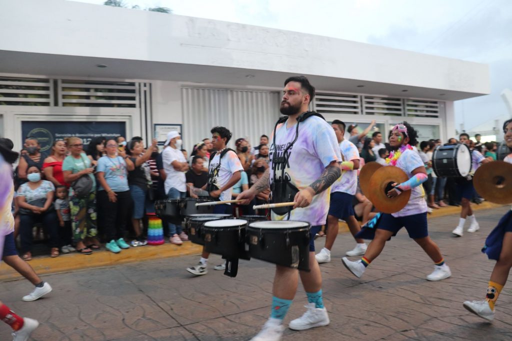 More members of the marching drum corps wearing pastel tie dye shirts. Seven members are visible and six have drums while one has a large pair of cymbals. They are marching in one of the Cozumel Carnaval parades in daylight down a spectator lined street with a white building in the background.