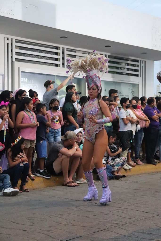 A young woman entertainer walking the parade route for the Cozumel Carnaval parade. It is still daylight and there is a white building in the background and spectators lining the street. She is wearing an ornate lavender leotard costume with lavender knee high boots, lavender arm bands and a tall feathered headpiece.