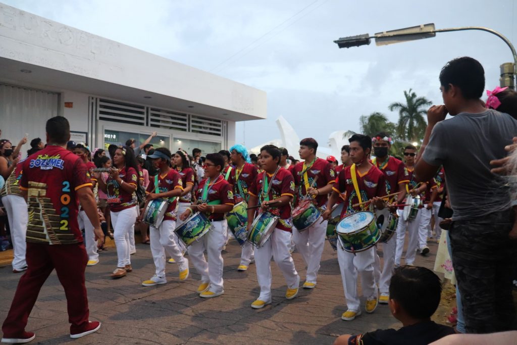 A drum corps marching in the Cozumel Carnaval parade. A dozen members are shown marching in rows with red shirts and white slacks, playing drums that they wear strapped to them. The band leader is walking backwards in front of them while giving directions. It is daylight still and there is a white building in the background. They are all wearing matching bright yellow athletic shoes.