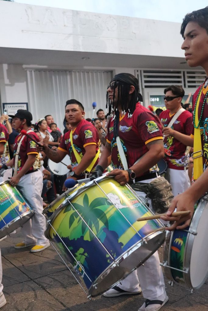 A drum corps marches in the Cozumel Carnaval parade. The young men are wearing red shirts with insignia, white slacks and carrying large drums in front of them. The drums have an island print graphic in greens and blues.