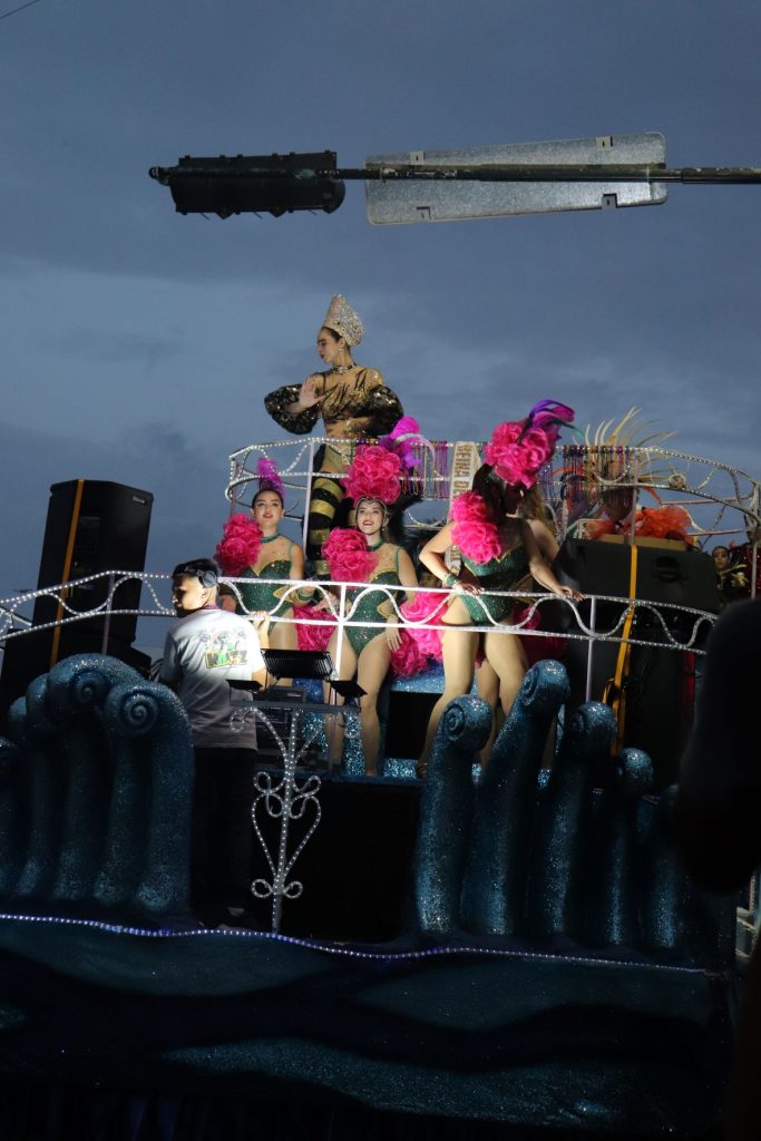 A full height left front view of a parade float at Cozumel Carnaval. The upper level has a queen with an ornate costume and crown and the lower level has 3 women entertainers with hot pink feather headpieces and costumes with fluffy hot pink over a shoulder. The sky is dusky and the float is under a street sign/lamp.