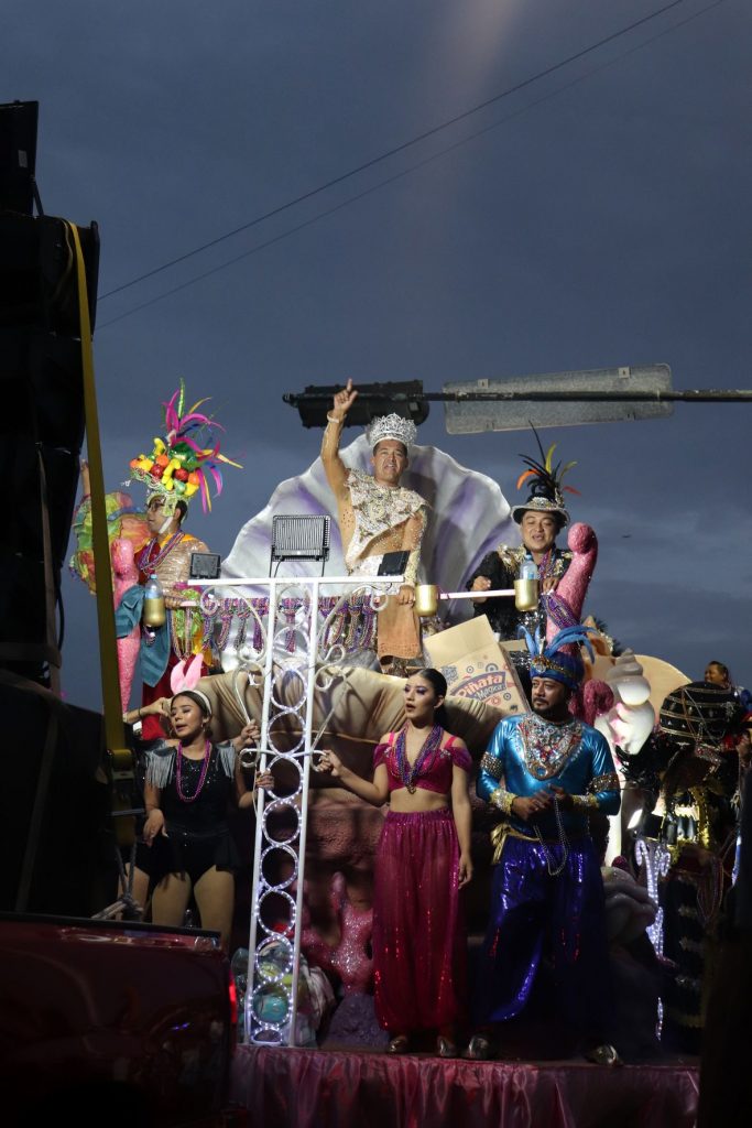 Frontal overview of a float at the Cozumel Carnaval parade. There is an upper level deigned as an open clam shell, with a man in a king costume waving to the crowds. The lower level has several performers in a variety of colorful costumes.