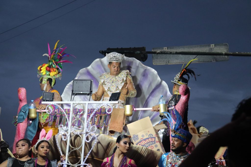 Cozumel Carnaval parade float with a man wearing sleeveless costume and crown on upper level. Upper level is designed like an open clam shell. The lower level has several other entertainers in various costumes. The sky is beginning to get dark and the float is rolling under an overhead street sign.