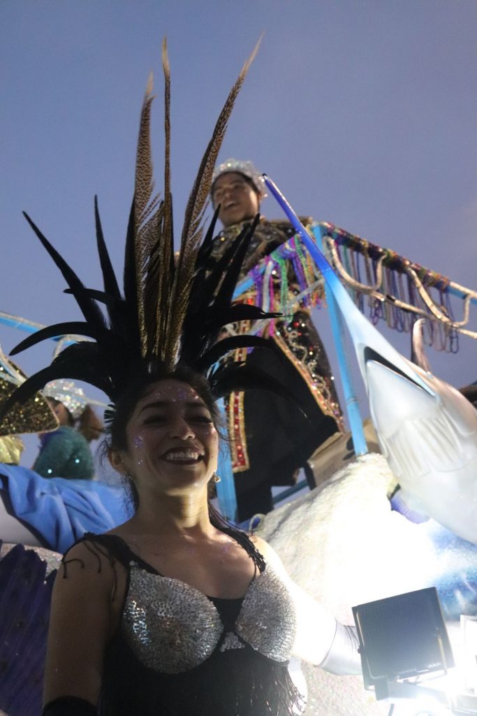 The sky is getting dark as this float rolls down the street at Cozumel Carnaval parade. A young latino man is standing on the upper level wearing a crown and a knee length royalty robe. A woman is in the forefront on the lower level of the float wearing a silver sequin bra top with a spikey feather headband.