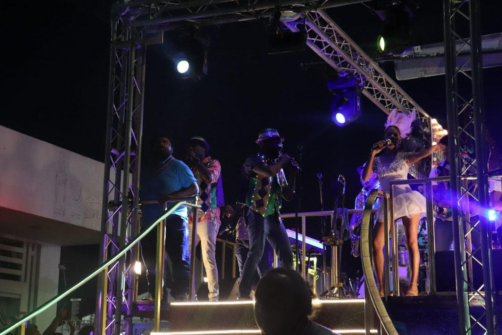 A group of dancers wearing bright Carnaval attire dance the night away on stage as the crowd watches. One woman wears a pink dress with matching feathers while a man wears a small hat and Mardi Gras colored clothing.